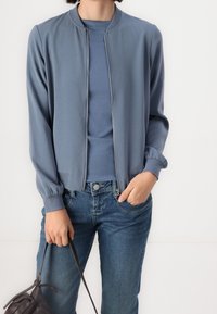 Lättvikts bomberjacka med blixtlås fram i dämpad blå färg, med ribbade ärm- och kragekanter. Matchad med en topp i samma färg och jeans i denim.