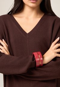 Suéter de punto burdeos con un escote en V profundo, combinado con una pulsera roja con un detalle dorado. Se destaca la textura suave y el ajuste relajado.