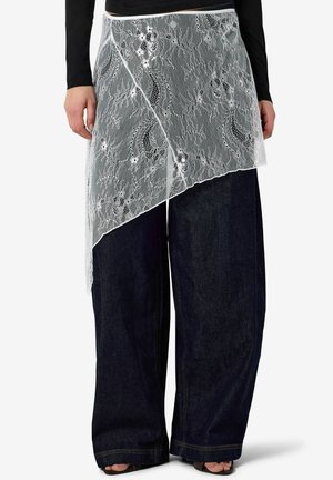 Nő aszimmetrikus fehér virágos csipkés átkötős szoknyában, sötét széles szárú farmernadrág fölött, fekete hosszú ujjú felsővel és nyitott orrú cipővel.