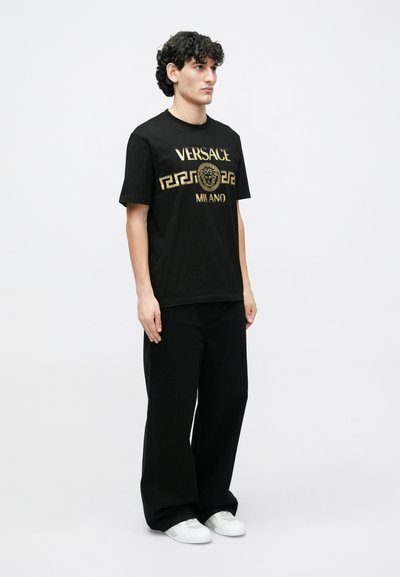 Černé tričko s krátkým rukávem z bavlny s zlatým nápisem "VERSACE" a "MILANO" a grafikou lva, kombinované s volnými černými kalhotami a bílými teniskami.