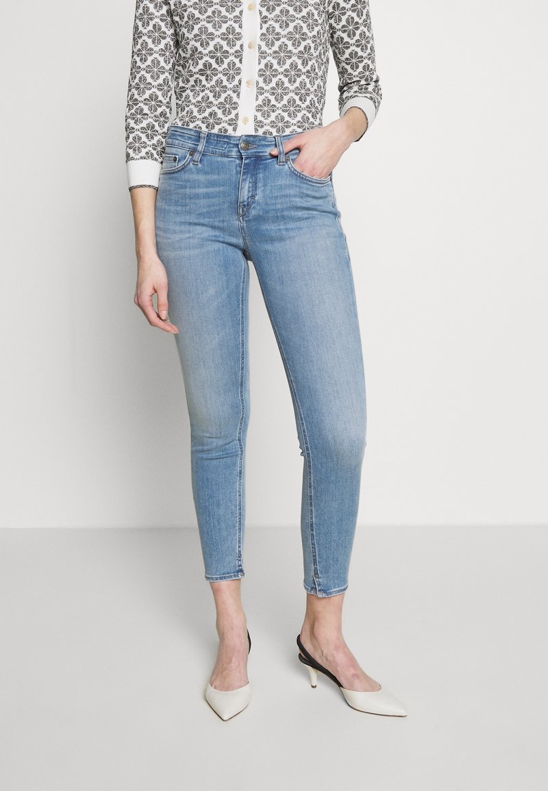 Jeans de denim azul claro con un corte slim, diseño de cinco bolsillos y un ligero desgaste. Combinados con una blusa de manga larga con estampado y tacones blancos.