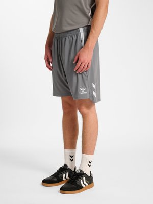 Mężczyzna ubrany w szare sportowe szorty hummel, białe skarpety z czarnymi chevronami oraz czarne trampki z białymi pasami w kształcie chevronów, stojący na białym tle.