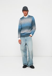 Pull bleu dégradé avec des tons gris, associé à un pantalon en jean large bleu clair et des chaussures noires. Comprend une casquette noire et une chemise blanche en dessous.