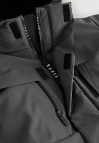 Grijze buitenkleding met een strakke vormgeving, voorzien van ritsen, klittenbands accenten en merknamen. Het materiaal lijkt glad en duurzaam.