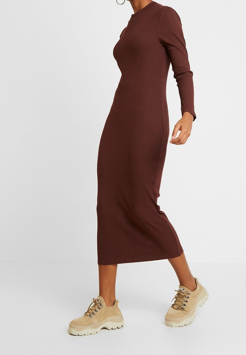 Personne portant une robe midi marron côtelée à manches longues, associée à des baskets beige épaisses, debout devant un fond uni.