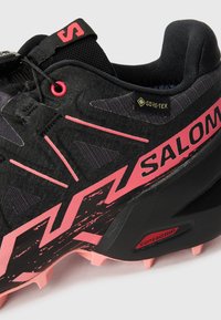 Chaussure de trail noire et rose avec une tige texturée, une étiquette Gore-Tex et une semelle extérieure Contagrip. Dispose d'un profil de crampons agressif et d'éléments de design distinctifs.