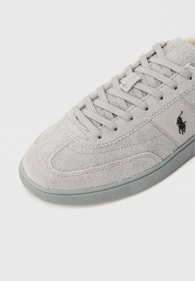 Γκρι σουέντ αθλητικό παπούτσι με στρογγυλεμένη μύτη, υφασμένη επιφάνεια, επίπεδα γκρίζα κορδόνια και ένα μικρό μαύρο λογότυπο στο πλάι. Λαστιχένια σόλα με αντιολισθητικό μοτίβο.