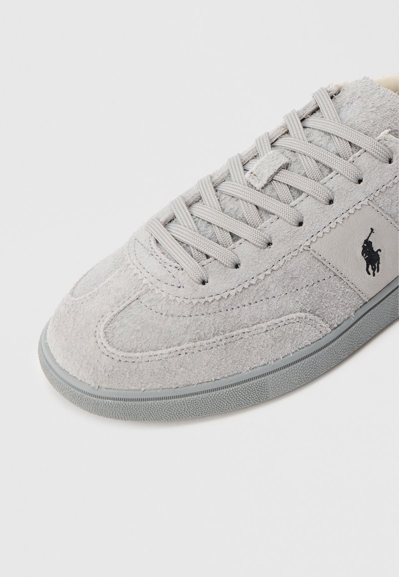 Sneaker in suede grigio con punta arrotondata, superficie texture, lacci piatti grigi e un piccolo logo nero sul lato. Suola in gomma con motivo antiscivolo.