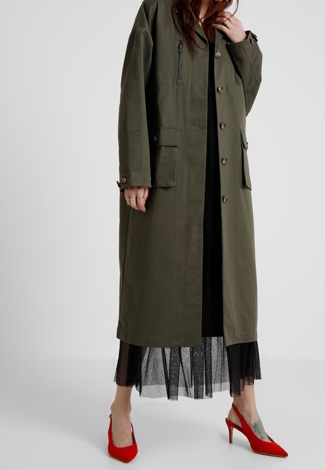 Femme portant un long manteau vert olive sur une jupe en résille noire et des chaussures à talons rouges, debout sur un fond clair uni.