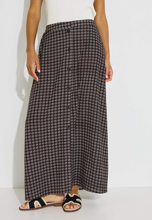 Mujer con una falda larga negra con estampado y botones delanteros, top blanco sin mangas y sandalias deslizantes negras y beige frente a una pared lisa.