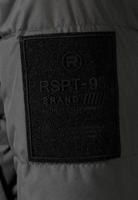 Pelēka auduma jaka ar lielu melnu teksturētu uzlīmi, kurā ir iegravēts uzraksts "RSPT-96 BRAND" un citi zīmola elementi.