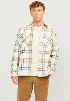 Plaid overshirt in white, beige, and brown with two chest pockets, button closure, and long sleeves; worn over a light-colored T-shirt.