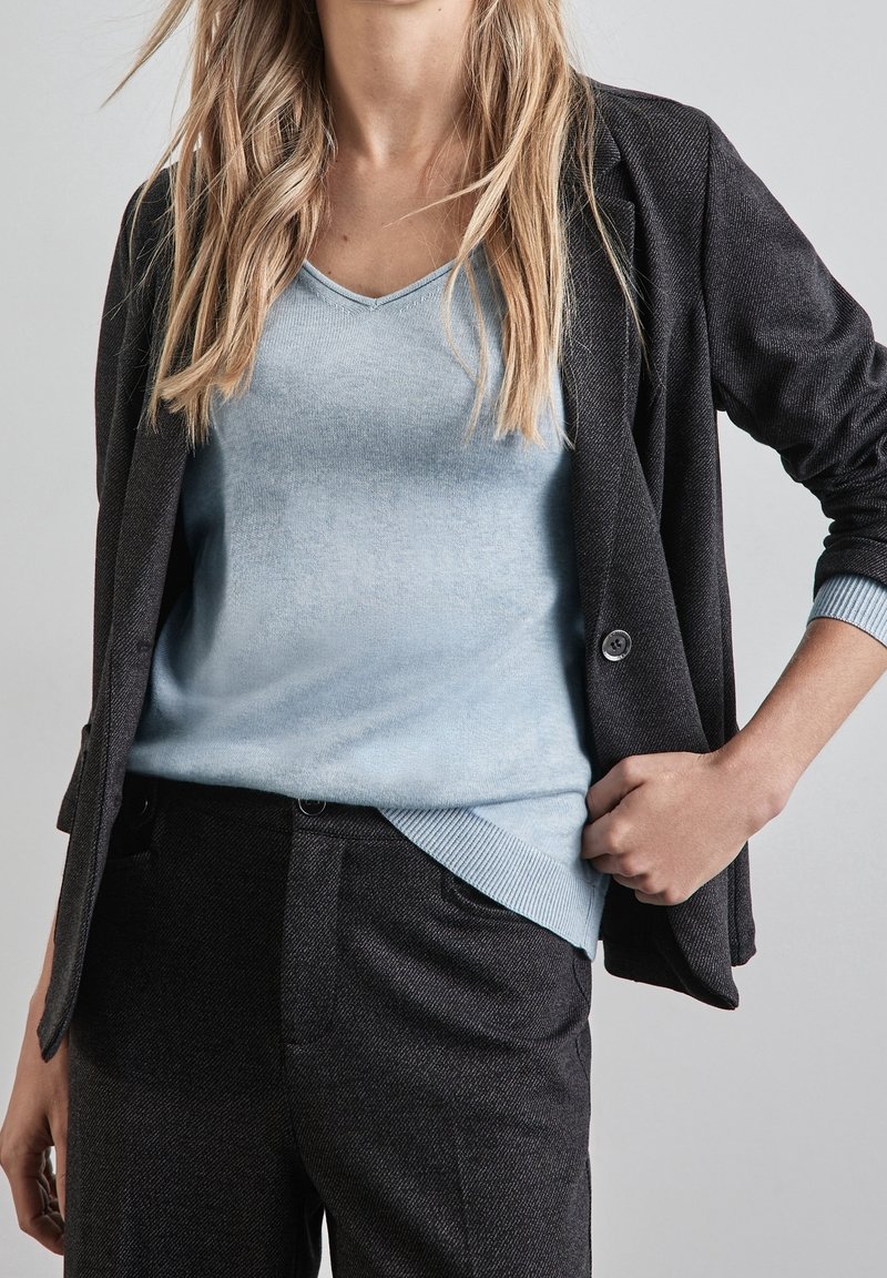 Femme portant un pull bleu clair à col en V sous un blazer gris foncé et un pantalon gris foncé assorti, debout avec une main sur la hanche.