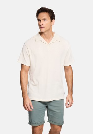 Hombre vistiendo una camisa polo color crema de manga corta y pantalones cortos grises remangados, posando contra un fondo liso y claro.