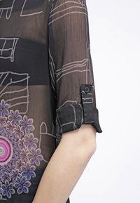 Blouse noire transparente avec des lignes géométriques grises et un détail de broderie florale colorée en bas. Présente des poignets roulés avec une fermeture à bouton.