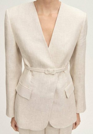 Blazer beige sur mesure avec col en V profond, ceinture fine assortie, épaules matelassées, poches à rabat et manches longues porté par une personne.