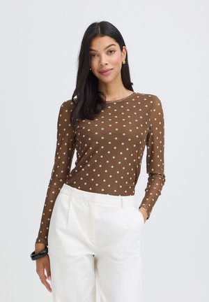 Kobieta o prostych czarnych włosach, ubrana w brązową bluzkę w grochy z długim rękawem, wpuszczoną w białe spodnie z wysokim stanem, z jedną dłonią w kieszeni.