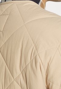 Beige gesteppte Jacke mit diagonalem Muster, leichtem Stoff und einem schwarz-weißen gemusterten Kragen. Nahtdetails sind sichtbar.
