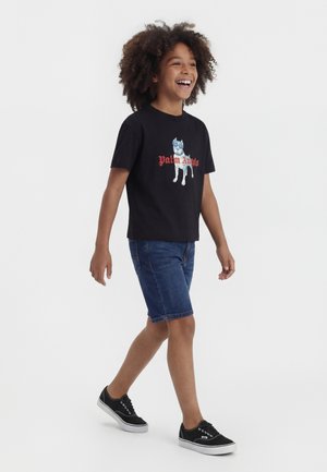 Vaikas su garbanotais plaukais šypsosi ir vaikšto, dėvi juodą marškinėlį su šuns grafika, džinsinius šortus ir juodus sportbačius su baltais padais.