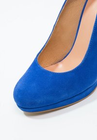 Modrá semišová bota na vysokém podpatku s kulatou špičkou a vykrojeným designem, s hladkým interiérem a pevným světle barevným podešvem.