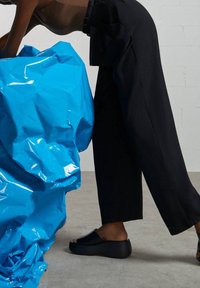 Une personne portant un pantalon en large évasé noir et des sandales à plateforme noires, penchée sur un grand objet en plastique bleu froissé sur un sol gris.