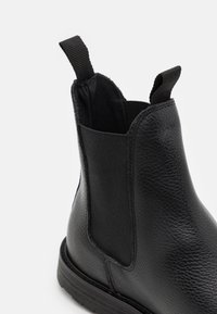 Botín Chelsea de cuero negro con paneles laterales elásticos, acabado texturizado y tira para facilitar el calzado. La suela es plana y duradera, diseñada para el confort.