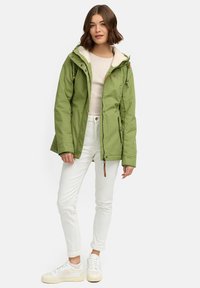 Grön vattentät jacka med en beige fleecefodrad huva, framdragkedja och sidofickor, ihop med vita jeans och vita sneakers.