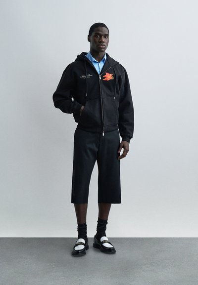 Άνδρας που φοράει μαύρο φούτερ με φερμουάρ και σχέδιο κόκκινου πουλιού, μπλε πουκάμισο, μαύρο ευρύ κομμένο παντελόνι, μαύρες κάλτσες και μαύρα-λευκά loafers, στέκεται μέσα σε εσωτερικό χώρο.