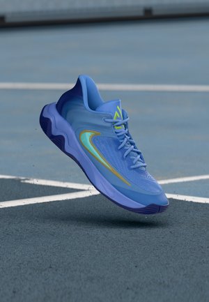 Chaussure de sport bleue avec un design swoosh vert et jaune flottant au-dessus d'un terrain de sport gris avec des lignes blanches.