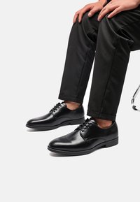 Scarpe da uomo in pelle nera con finitura liscia, punta tonda e design con lacci, abbinate a pantaloni neri, che mostrano uno stile pulito e classico.