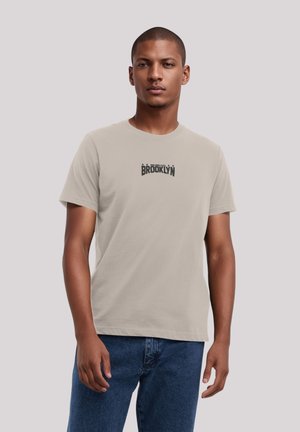 Beige Baumwoll-T-Shirt mit kurzen Ärmeln und runder Halslinie. Mit einem schwarzen Grafikdruck von "BROOKLYN", der mittig auf der Brust angebracht ist.
