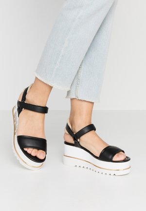 Czarne sandały na koturnie z paskami, z gładką, skórzaną cholewką, białą podeszwą na platformie i zygzakowatą, teksturowaną podeszwą zewnętrzną. Wysokość obcasa jest znaczna.