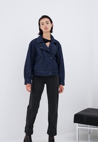 Mörk denimjacka med kort längd, bred krage, framknappar och två sidofickor, i kombination med svarta, uppvikta byxor och ankstövlar.
