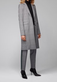 Abrigo largo de cuadros grises con solapas, bolsillos laterales y un corte ajustado, combinado con pantalones negros ajustados que tienen franjas verdes en los lados y botas de tacón negras.