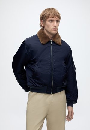 Man draagt een marineblauwe bomberjack met een bruine shearlingkraag en beige broek, staand tegen een effen lichte achtergrond.