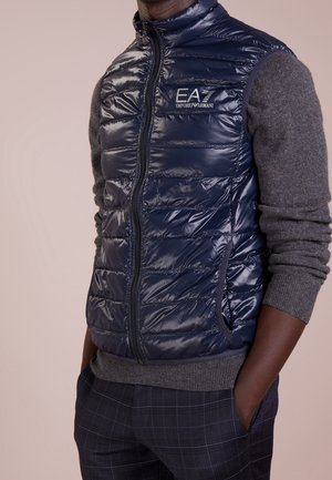Homme portant une veste matelassée bleu marine avec le logo EA7 Emporio Armani, manches de pull gris, et pantalon à carreaux foncé, mains dans les poches.