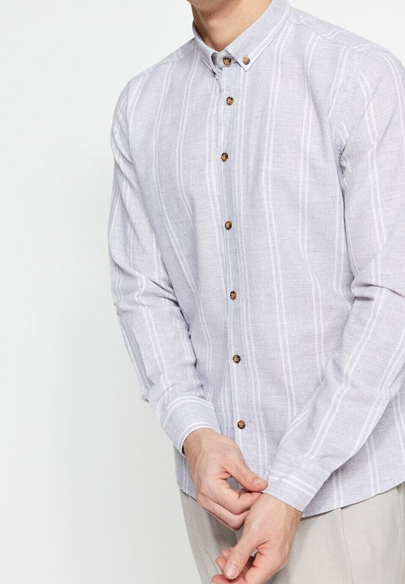 Homme portant une chemise boutonnée gris clair avec des rayures blanches verticales, en train de fermer le poignet, visible du cou aux hanches sur un fond blanc.
