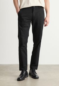 Pantaloni neri con una texture liscia, caratterizzati da un taglio dritto e tasche anteriori. Abbinati a eleganti scarpe nere, su uno sfondo neutro.