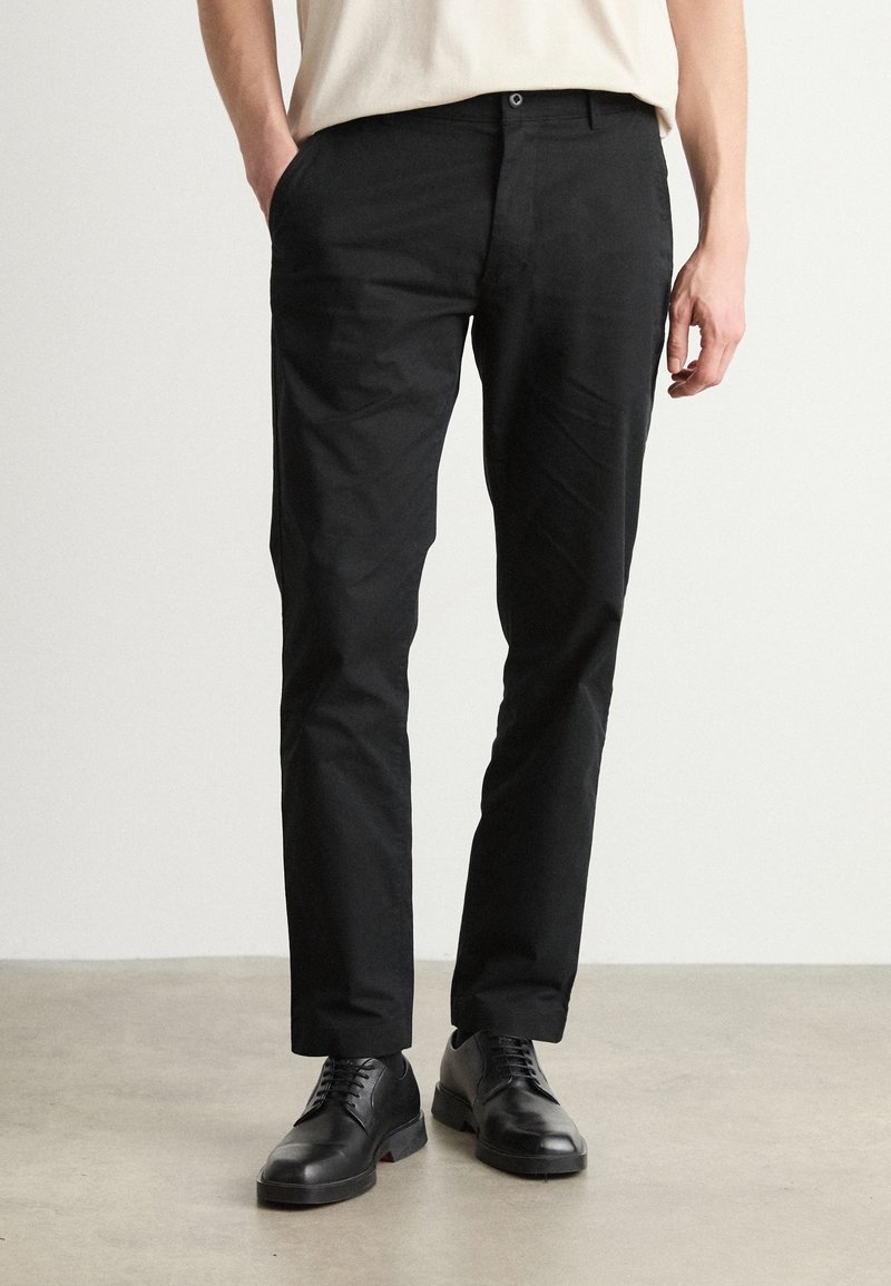 Pantaloni neri con una texture liscia, caratterizzati da un taglio dritto e tasche anteriori. Abbinati a eleganti scarpe nere, su uno sfondo neutro.