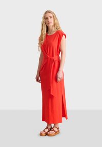 Robe rouge, longueur genou, avec manches courtes et taille nouée. Fabriquée en tissu doux, elle présente une fente sur le côté pour faciliter le mouvement. Portée avec des sandales marron.