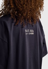 Svart bomullst-shirt med tryckt text "F.U.T. PHYSICAL EDUCATION & TRAINING" och "B-583" på baksidan, med avslappnad passform och mjuk textur.