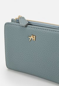 Portafoglio in pelle blu con texture, hardware dorato e logo. Presenta una chiusura con zip e una foderatura interna liscia. Forma rettangolare compatta.