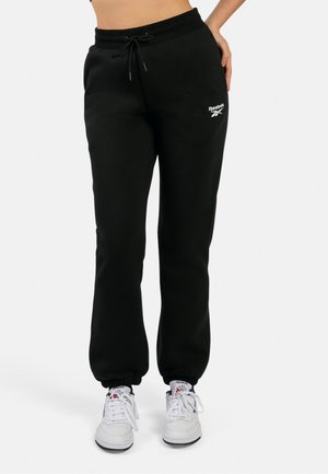 Persona con pantalones jogger negros de Reebok con cintura de cordón y zapatillas blancas de Reebok, de pie sobre un fondo blanco.