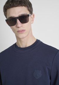 Sudadera navy con un emblema de oso en relieve en el pecho. Modelo usando gafas de sol oscuras, con una textura de tela suave y cuello redondo.