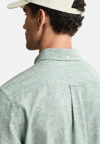 Grünes Leinenhemd mit einem Button-down-Kragen, verfügt über eine Rückenpasse und texturierten Stoff. Das Hemd wird mit einer hellen Kappe getragen.