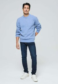 Pull en maille bleu clair avec col rond, associé à un jean foncé et des baskets blanches. Le mannequin se tient avec une main dans la poche.