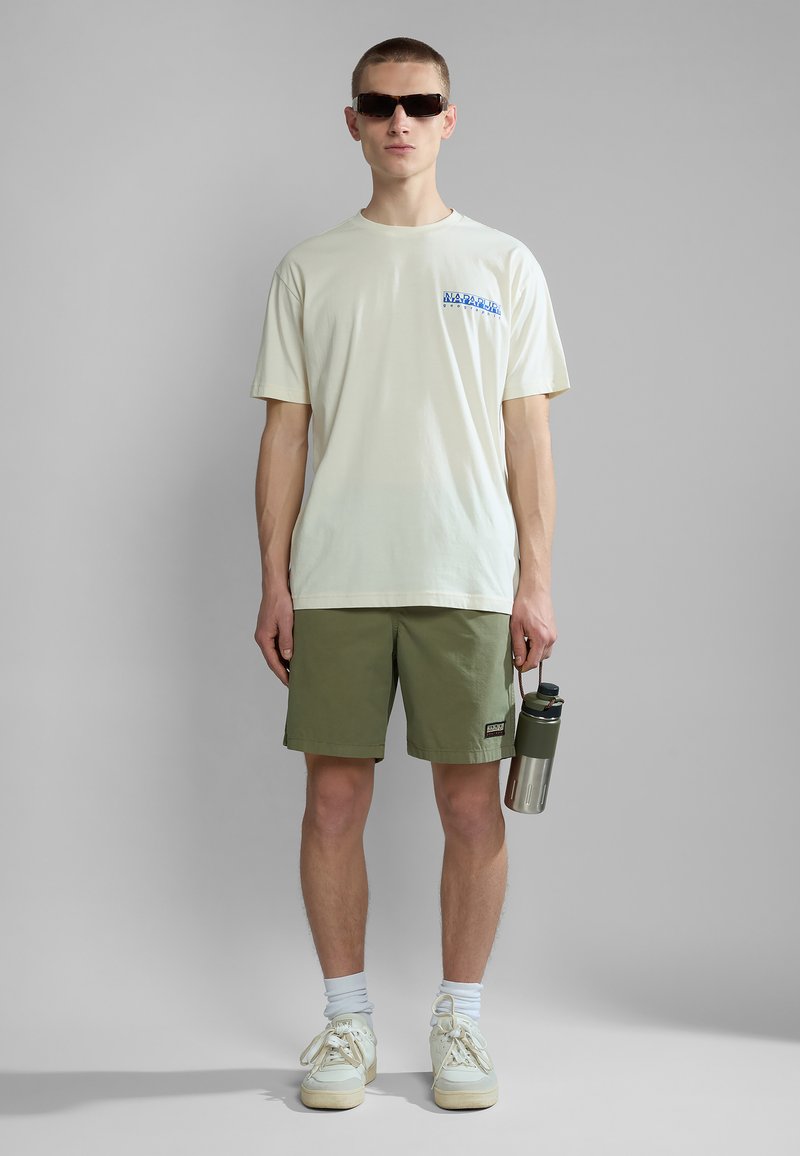 Young man wearing sunglasses, white T-shirt, olive shorts, white socks, and sneakers, holding a green and silver water bottle by his side.