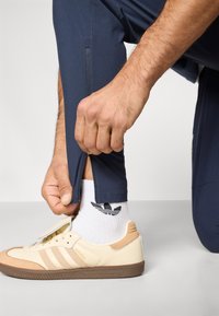 Elegantne mornarsko plave sportske hlače sa bočnim patentnim zatvaračem; bež tenisice s tri pruge i teksturiranim gumenim potplatom; bijele čarape s logotipom.