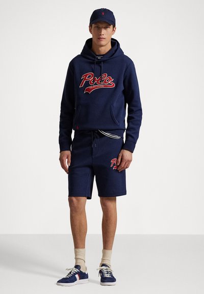 Sudadera de color azul marino con el logo rojo de "Polo", acompañada de pantalones cortos a juego. Tela ligera, bolsillo tipo canguro y cintura con rayas en contraste.