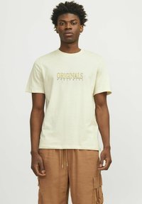 Hombre con cabello corto y rizado, vistiendo una camiseta beige con el texto "ORIGINALS WORLDWIDE" y pantalones cortos de cargo marrones, de pie contra un fondo liso.